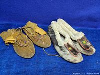 Two pairs of antique moccasins placed side by side on blue fabric background, showing one pair with spotted fur and fur collar, the other brown suede pair with leather tie.
