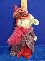 Full view of Bearington plush teddy bear with 1850s style clothing, including floral-patterned purple coat and red skirt, with large decorated fedora hat and pink feather boa. Measuring ruler in background shows height approximately 14 inches.
