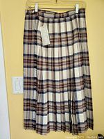 Full view of new with tags Aljean pleated tartan wool skirt, size 8, hanging on a yellow wall.