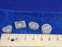 Four small pewter trinket boxes lined up next to a ruler showing their size around 1.25 to 2 inches each