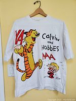 Front view of the white T-shirt with colorful graphic of Calvin and Hobbes characters and YA, AAA lettering.