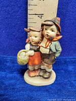 Front view of vintage Goebel Hummel figurine showing two children, a boy and a girl, against a blue background with measuring scale showing about 4 inches tall.
