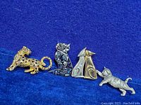Full view of all five cat and leopard brooches on blue velvet background, showing variety of styles and materials.