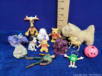 Collection of 13 assorted collectible toy figures including Winnie the Pooh, plush dog, M&M keychain, and others shown with ruler for scale