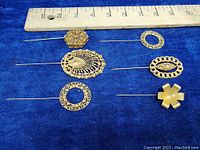 Six gold-tone filigree hat and scarf pins arranged on blue fabric with measuring ruler showing size.