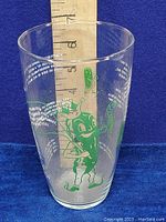 Full view of the vintage glass cup with ruler showing height, featuring green anthropomorphic dill pickle character and white nursery rhyme text.