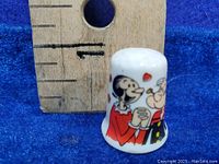 Close-up image of thimble with Popeye and Olive Oyl characters next to a ruler showing height about 2 cm.