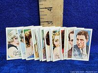 Stack of cigarette cards showing painted portraits of various 1930s film stars with a wooden ruler for scale indicating approximately 2 inch height.