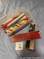 Plastic bin containing several plain taper candles in various colors, yellow spiral candle with red/black design, and loose black taper candles beside the bin. Red taper candles in sealed pack in front.