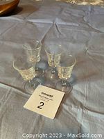 Four clear cordial glasses with decorative etched pattern arranged on a fabric surface along with a MaxSold lot number card.