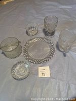Photo showing five clear glass decorative table items on light blue fabric background: a small pitcher, an ice bucket, a round plate, and two glasses with intricate pressed glass patterns.