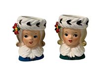 Front view of both ceramic toothpick holders showing detailed female faces and white fur-trimmed hats with red flower accent.