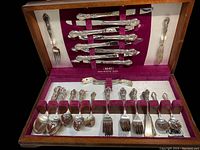 Overview of the entire silverware set in a wooden case with burgundy felt lining showing knives at the top and forks and spoons at the bottom.