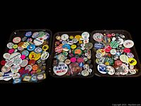 Three trays filled with various vintage pins of political, regional, and humorous nature, showing wide variety and quantity.