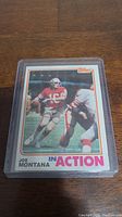 Photo of Joe Montana football card from the second year, encased in a protective plastic sleeve showing the front of the card with Joe Montana in red and gold uniform on the field.