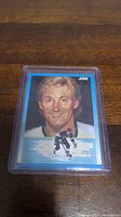 Photo of the Wayne Gretzky hockey card in a protective plastic holder, showing the front illustrated portrait of Gretzky with a blue frame and hockey skating image.