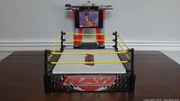 Photo of WWE RAW wrestling ring with white mat and three yellow ropes, missing the bottom rope, sitting on a wooden surface.