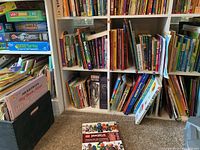 Wide view of five shelves of assorted children's books including boxed board games and puzzles on left side and a black storage box beneath the shelves. Books vary in size and colors.
