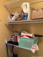 Overall view of cleaning supplies on wire shelves including large bottle of Scotchgard fabric protector, cleaning cloths and pot holders in green basket, and spray bottles below.