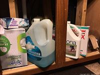 Various cleaning supplies arranged on a wooden shelf including Swiffer small trash bags box, Windex large jug, Bona floor cleaner box