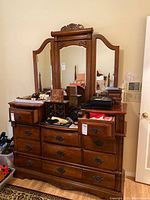 Front view of brown wooden dresser with eleven drawers and three-piece mirror on top, displaying surface clutter and decorative handles.