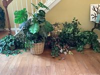 Several faux plants including a large leafy plant in a woven basket, various trailing and climbing faux plants in baskets with assorted green leaves, displayed on a hardwood floor.
