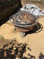 Full view of round metal fire pit with rust and lattice pattern, sitting on three legs outdoors on bare dirt.