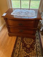 Pair of Stanley night stands positioned side by side on hardwood floor with area rug. Both have a brown wood finish, two drawers, curved fronts, and metal handles.