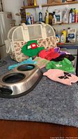 Photo showing very small colorful pet outfits displayed beside dual compartment feeding bowl and beige patterned pet carrying case on a counter in a garage.