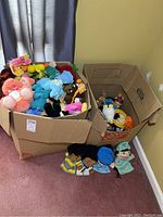 Two large cardboard boxes filled with various plush toys and hand puppets arranged on a carpeted floor.