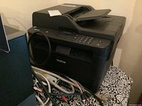 Side view of black Brother multifunction printer placed on a patterned surface with cables around it.