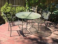 Full view of outdoor metal table with glass top and two metal chairs on wooden deck, sunlight casting shadows.