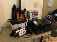 Overview of two consoles, two Kinect sensors, VR headset, guitar controller and stack of game cases on table