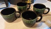 Four Blue Mountain Pottery mugs arranged showing rounded bodies, green and blue glaze with black handles and black interior.