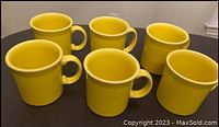 Six yellow Fiestaware mugs arranged on a black surface with cream rim detail visible.