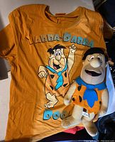 Front view of orange Flintstones T-shirt with Fred Flintstone graphic and plush doll of Fred Flintstone.