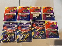 Top-down image of eight miniature vehicles, including six sealed on blister cards and two sets of three miniature cars packaged on cards.