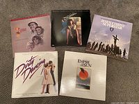 Photo of five movie soundtrack vinyl records laid out on a carpeted floor, showing album covers for 'Oscar', 'Flashdance', 'Jesus Christ Superstar', 'Dirty Dancing', and 'Empire of the Sun'.