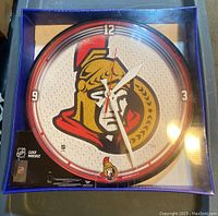 Full front view of the new and sealed Ottawa Senators wall clock in blue packaging, showing the clock face with team logo and clock hands.