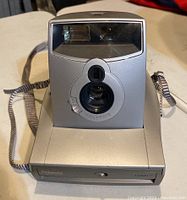 Front view of Polaroid 1200FF instant camera, showing lens, viewfinder, and flash unit with attached strap.