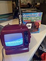 Portable black and white television with AM/FM radio powered on showing a black and white static screen, placed next to original product packaging box.