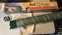 Photo showing the rolled green puzzle mat with Roll-O-Puzz branding, instruction sheet, a small circular container, and original box.