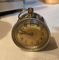 Front view of small vintage metal clock with yellowed dial, black numbers, and ornate center design