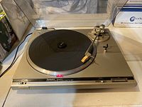 Full view of the Technics turntable powered on with the tonearm positioned to the side, on a surface with a dust cover propped open behind it.