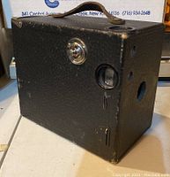 Overall front angle view of the vintage Buster Brown box camera showing the lens, shutter button, and leather handle.