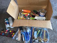 Box containing various paint supplies including numerous small paint bottles and spray paint cans, paintbrushes in multiple sizes and types, watercolor palette