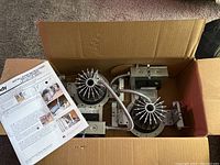 Open cardboard box showing multiple recessed light fixtures with fins for heat dissipation, mounted on adjustable metal brackets with flexible conduit wiring. Installation instructions are also visible inside the box.