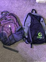 Photo showing two backpacks on a carpeted floor, one black and gray with Coop logo, the other purple and black.