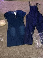 Two of the outfits including the jean dress shirt with skirt on the left and dark blue overalls on the right, both laying flat on carpet.