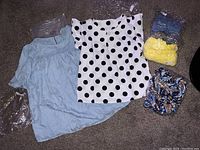 Photo showing five women's blouses, mostly still in plastic packaging. Light blue blouse, white with black polka dots blouse visible, several more folded blouses in packaging.